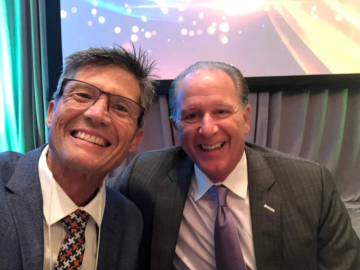 Podcast interview photo featuring Harold Earley in a gray pinstripe suit and purple tie, smiling beside a man in glasses.