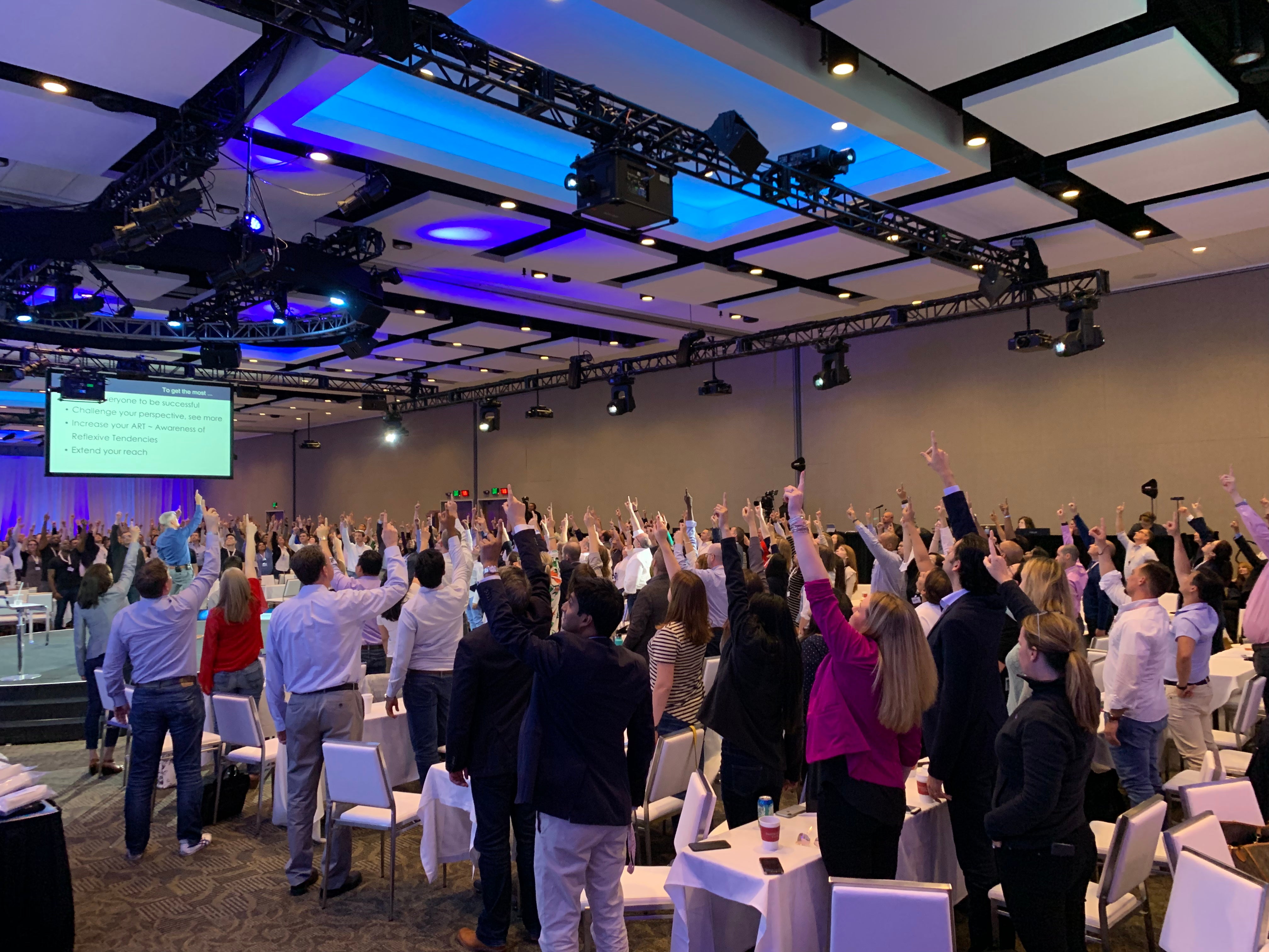 Large auditorium with people raising their hands, likely during a presentation or event.