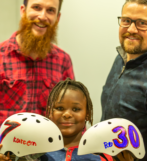 Two men standing behind a young child holding two white helmets with names on them.