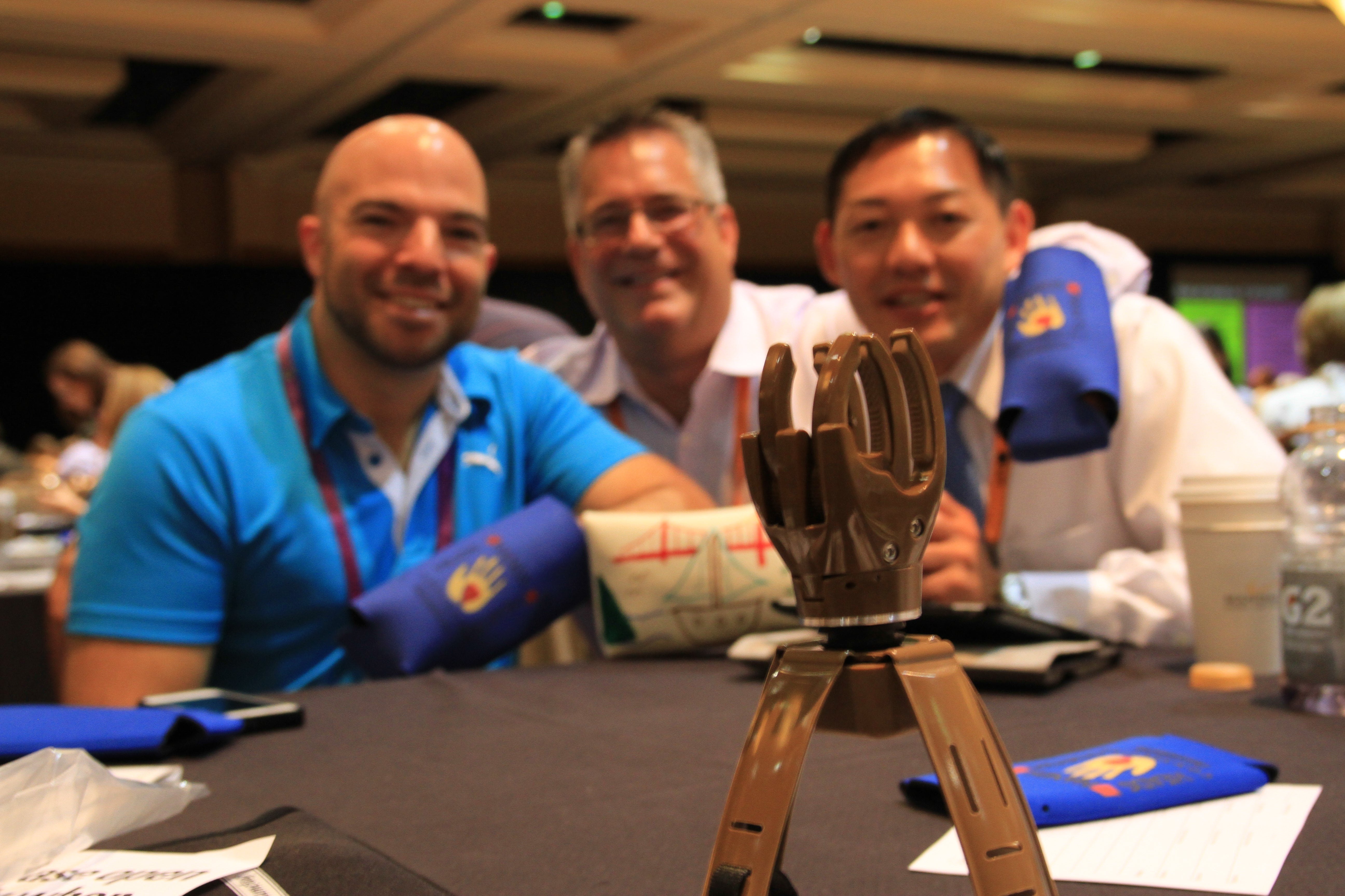 Three men sitting at a table with a prosthetic hand built from the Build-a-Hand teambuilding experience