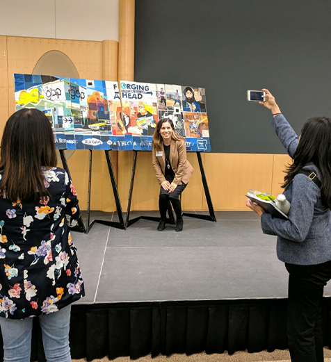 Woman taking a photo of another woman in front of a display board with various images and text.