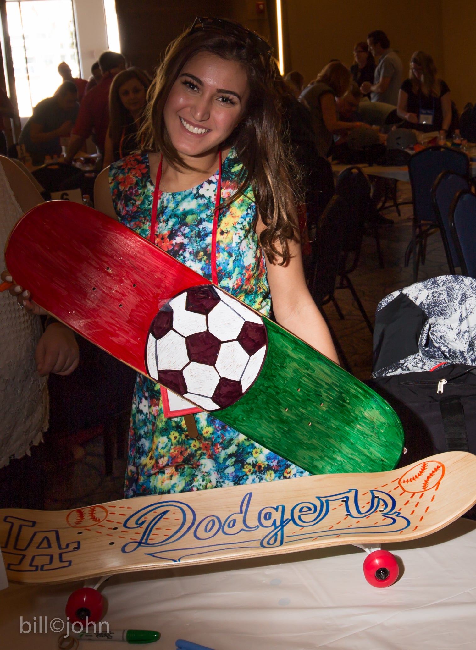 Woman holding a decorated skateboard with sports-themed design in a crowded indoor setting.