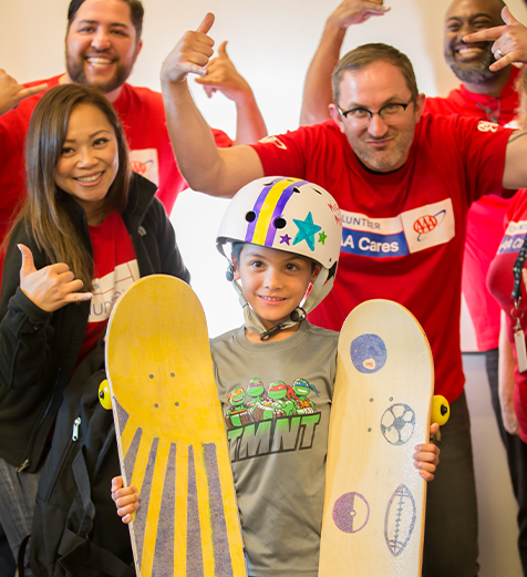 Child with a helmet and two skateboards, surrounded by adults in red shirts, likely at an event or program.