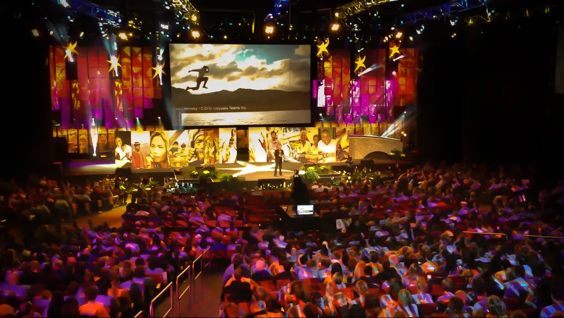 Large indoor event with stage, audience, and large screen displaying a scenic image.
