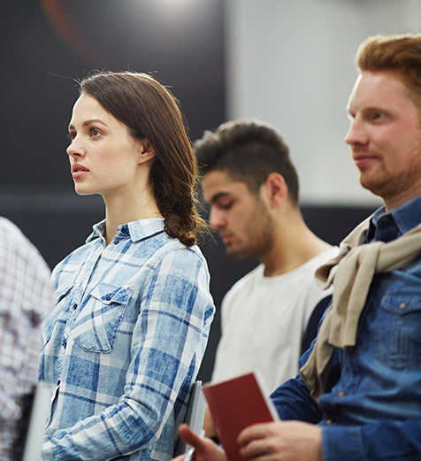 Group of people standing in a line, with a woman in a blue plaid shirt looking to the side.
