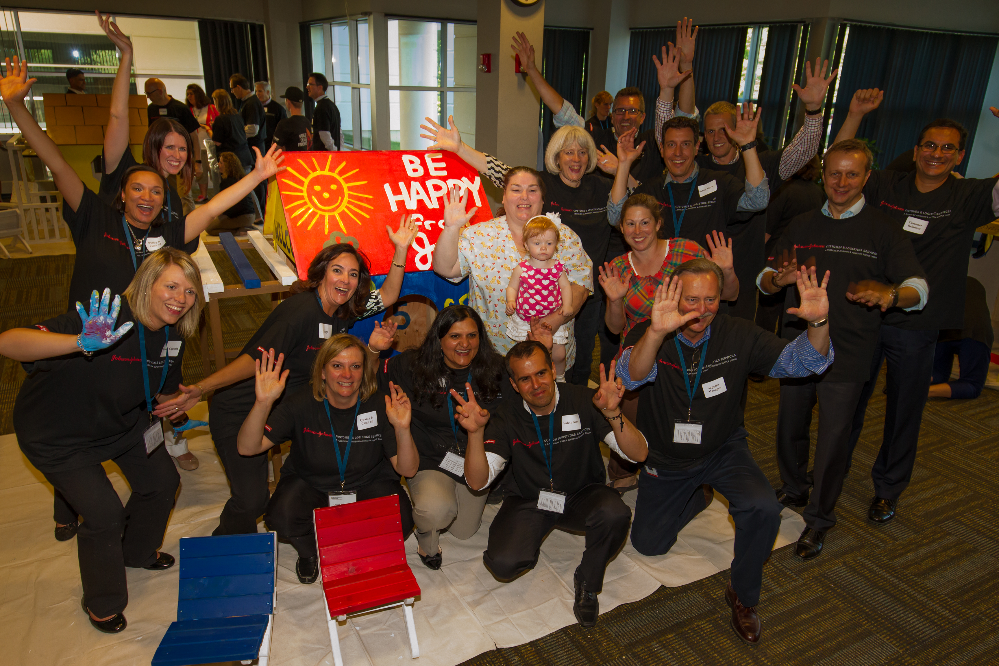 Group of people posing joyfully with a 'Be Happy' sign in an indoor setting.