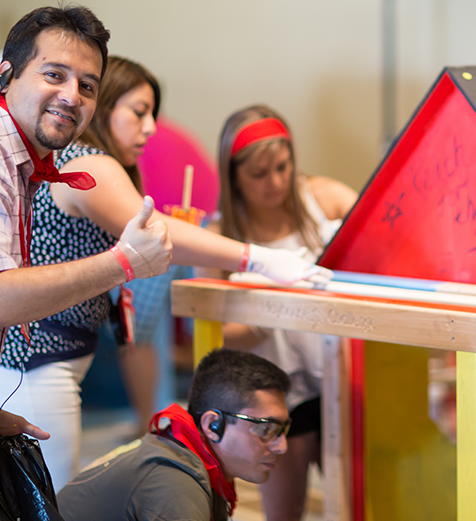 Person giving a thumbs up in front of a screen with a red triangle, surrounded by people in a casual setting.