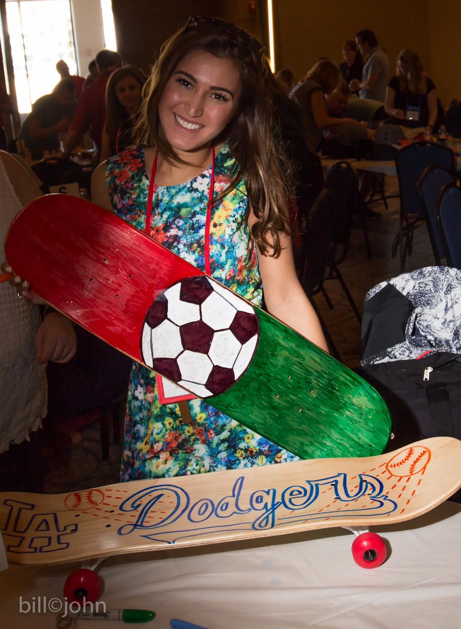 Woman holding a decorated skateboard with sports-themed design in an indoor setting.