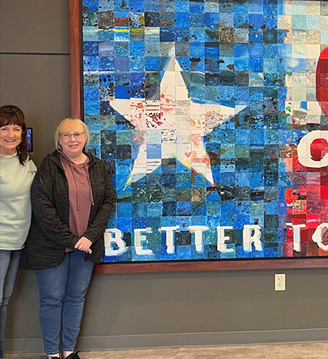Two women standing in front of a colorful mosaic wall art with a star and text.